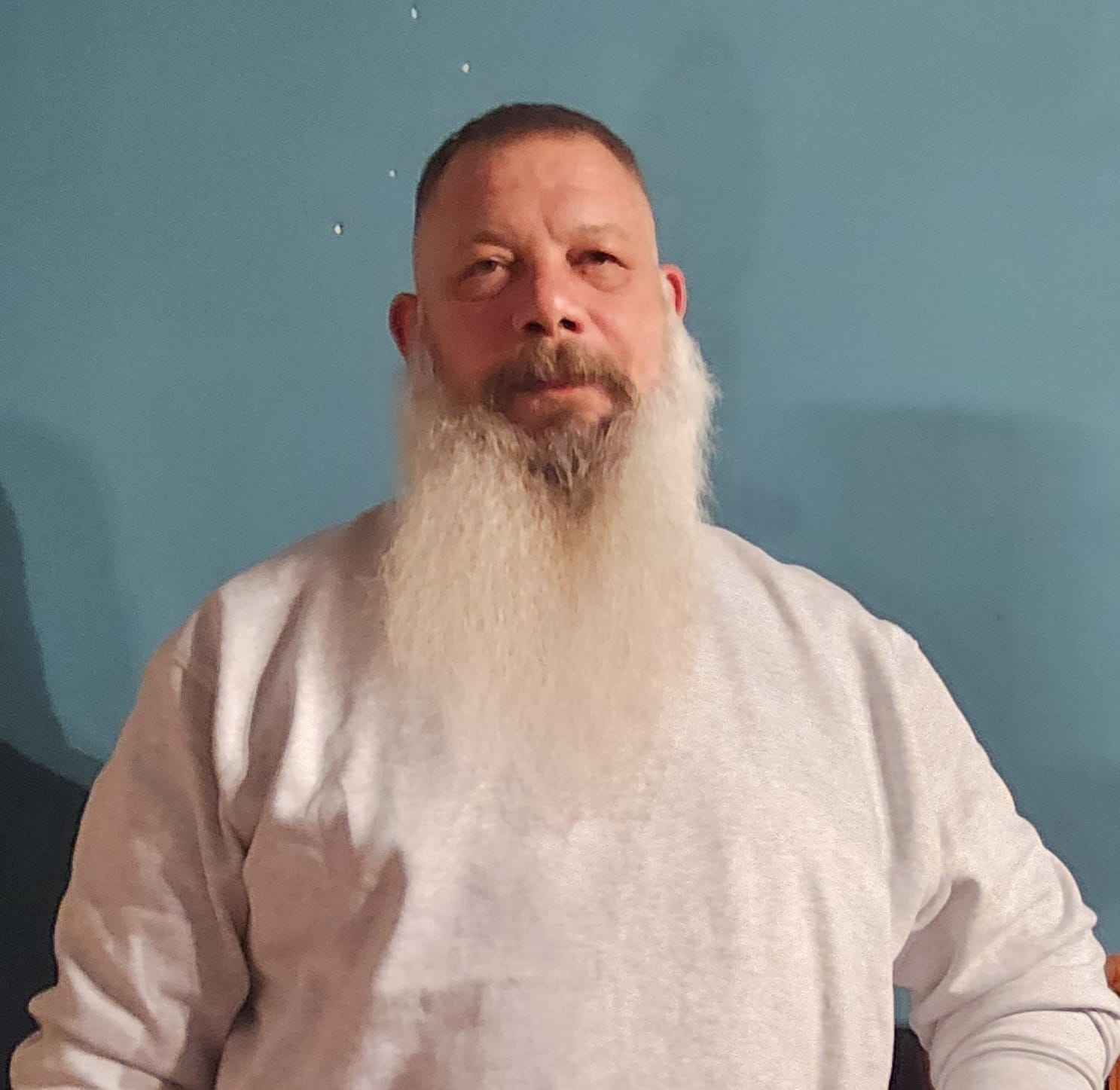 Unser Verein 21 Mann mit langem weißem Bart in einem grauen Langarmshirt vor einer blauen Wand – Mitglied des Bundes Deutscher Veteranen.